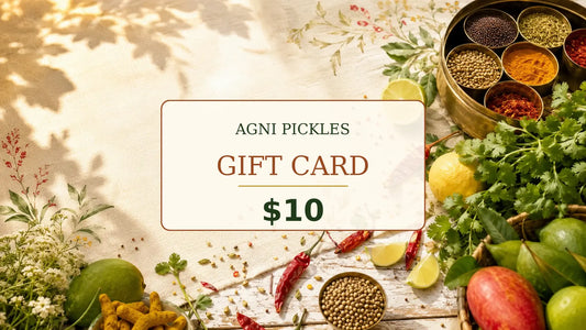 A promotional gift card for Agni Pickles, priced at $10, displayed on a rustic wooden table surrounded by fresh ingredients like limes, red chilies, coriander seeds, and spices.