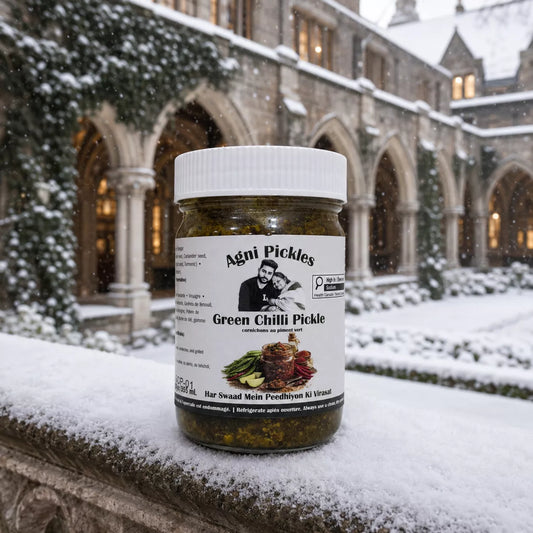 A jar of Agni Pickles Green Chilli Pickle sits on a snow-covered stone ledge in front of a historic stone building with snow on the roof and courtyard during a light snowfall.