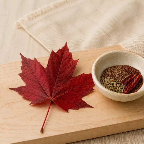 A dried red maple leaf and a small ceramic bowl containing brown seeds and red chili peppers, arranged on a light wood board with a cream fabric background.