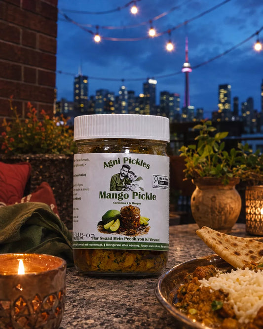 A jar of Agni Pickles Mango Pickle is displayed on a stone table in a restaurant setting with a city skyline at dusk in the background. A lit candle, a plate of food, and potted plants are visible around the jar.