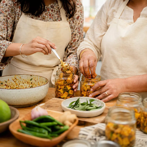 Two people wearing cream aprons are preparing food, with one spooning a chunky orange-yellow curry paste into glass jars while others prepare ingredients on a wooden table.