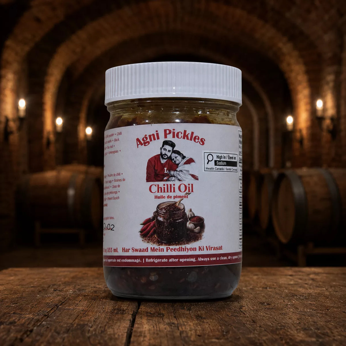A jar of Agni Pickles Chilli Oil placed on a rustic wooden table with a blurred background of an arched stone hall with warm lights.