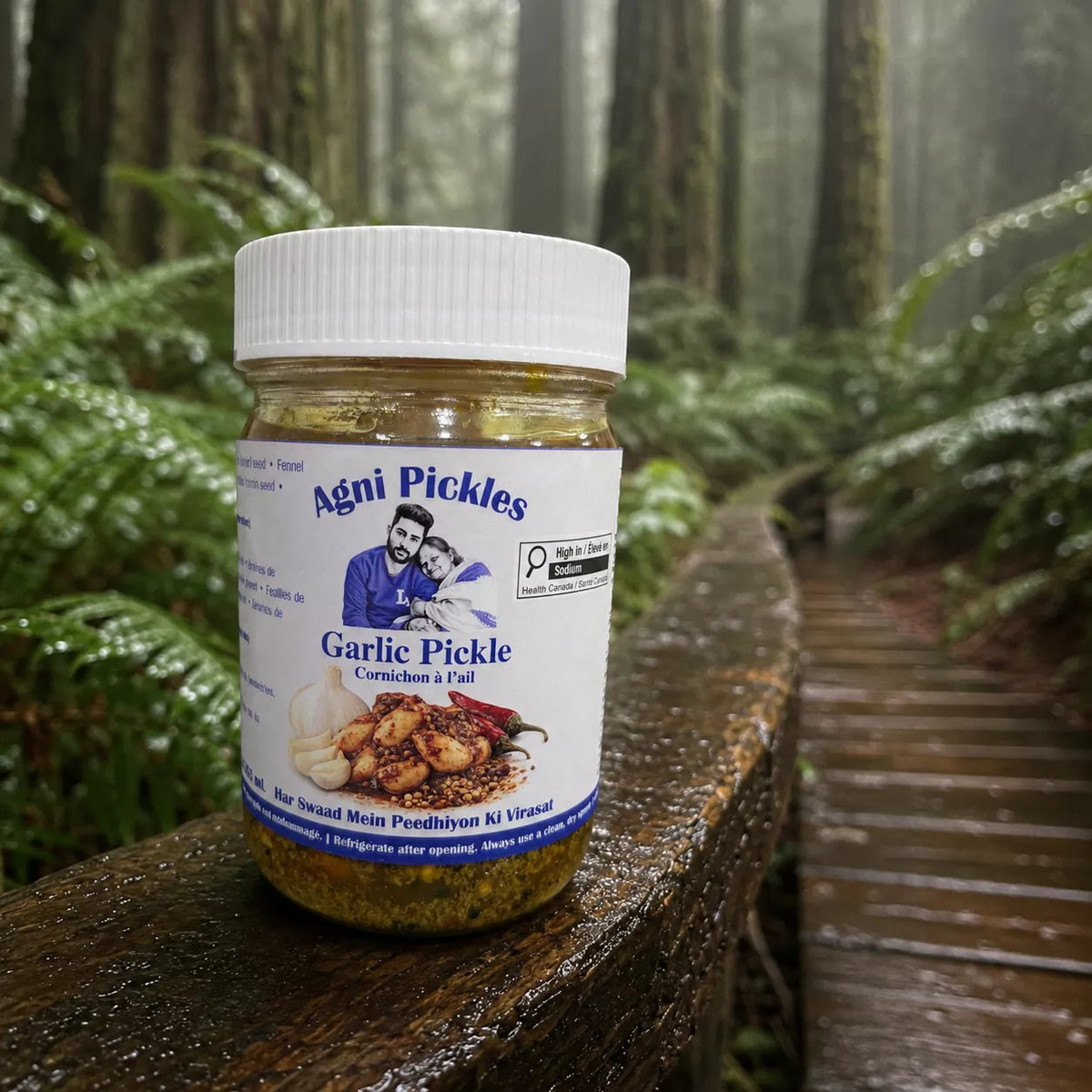 A jar of Agni Pickles Garlic Pickle sits on a wet wooden railing in a misty forest setting. The clear glass jar shows the golden-brown pickled contents, and the white label features the brand name 'Agni Pickles' and an illustration of a man and child.