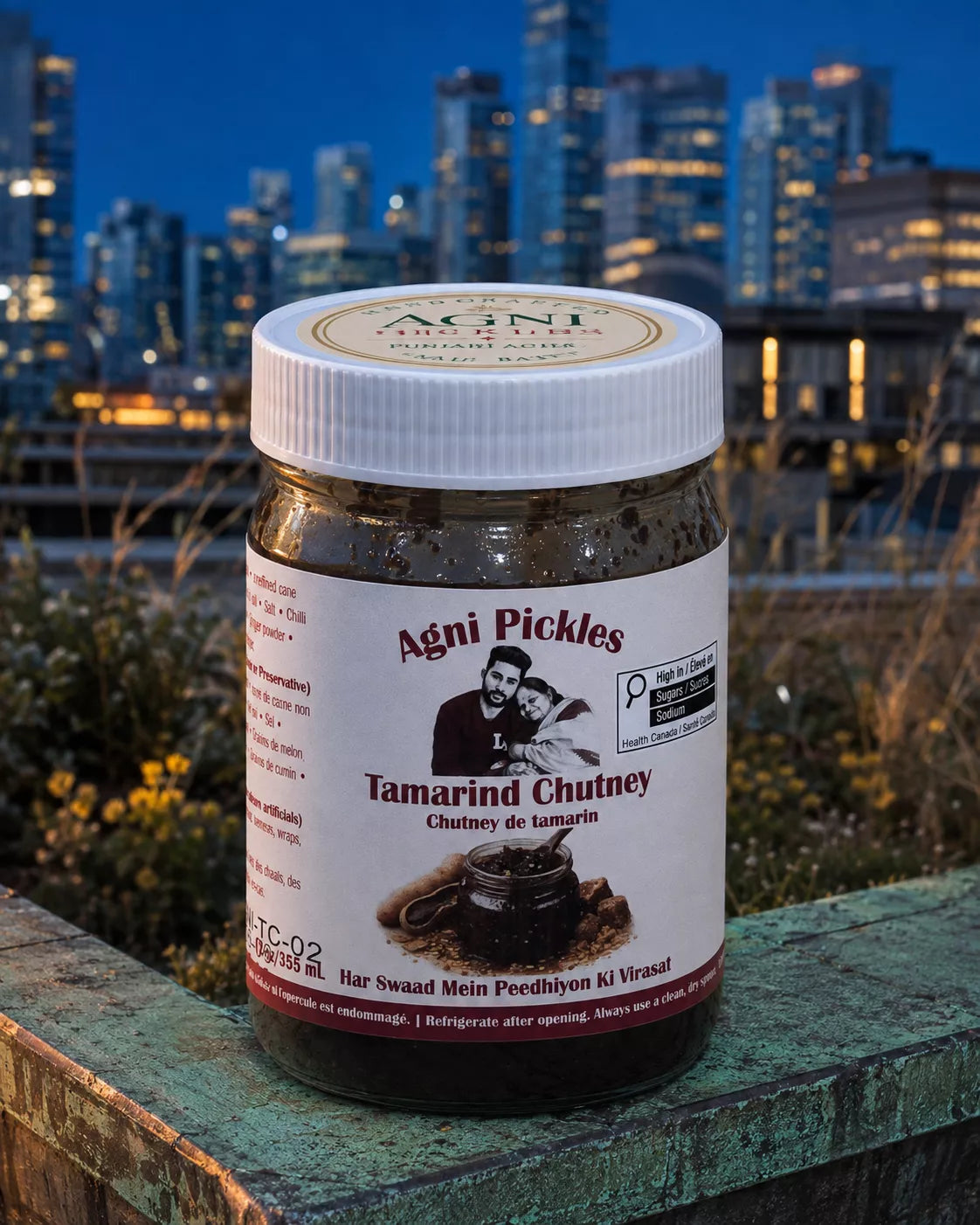 A jar of Agni Pickles Tamarind Chutney sits on a weathered green stone ledge with a blurred city skyline at dusk in the background. The jar has a white screw cap and a white label with red and black text.