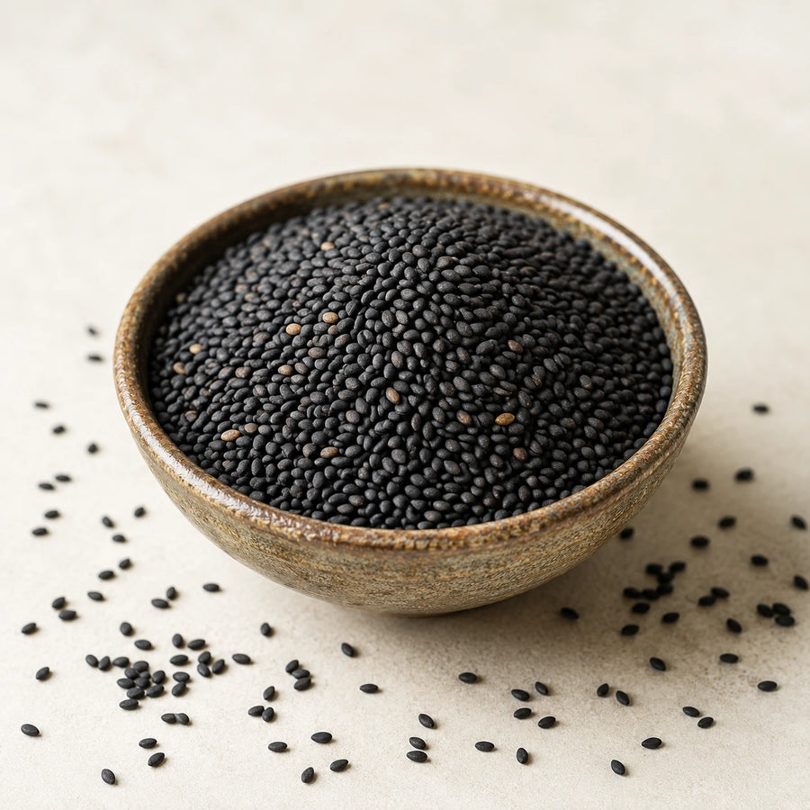Black sesame seeds piled in a small rustic ceramic bowl on a light beige surface, with a few scattered around the bowl.