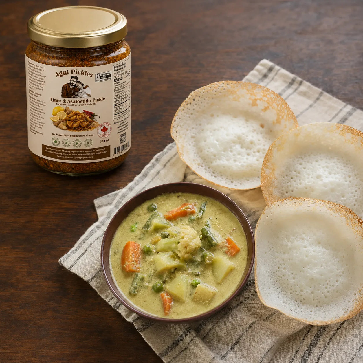 A jar of Mapi Pickles Lino & Anchoilla Pickle is shown next to a bowl of prepared pickle sauce with vegetables and three crispy round bread slices on a striped napkin over a wooden table.