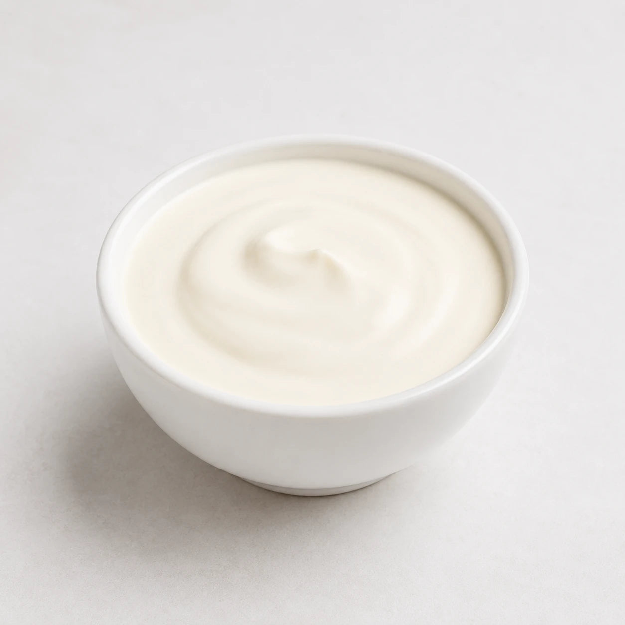 A small white ceramic bowl filled with creamy off-white yogurt, photographed from above on a light neutral background.