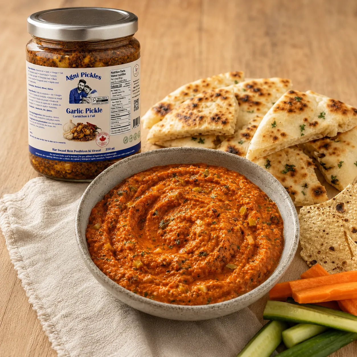 A bowl of orange-red garlic pickle dip in the center, with a jar of Ndai Pickles Garlic Pickle standing behind it and sliced pita bread, carrot sticks, and cucumber sticks arranged on a wooden surface.
