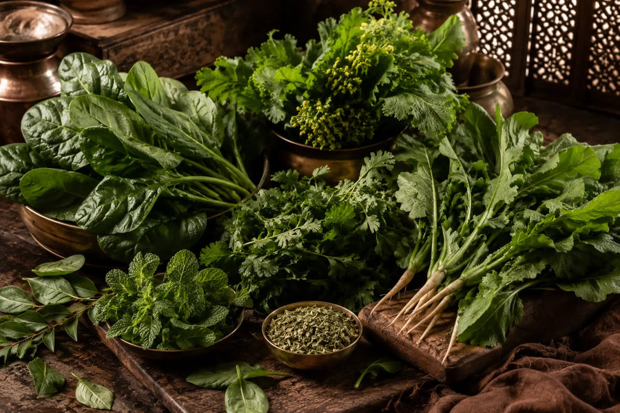A rustic arrangement of fresh green herbs and vegetables in brass bowls and on a wooden surface, with warm ambient lighting and a dark carved wooden background.