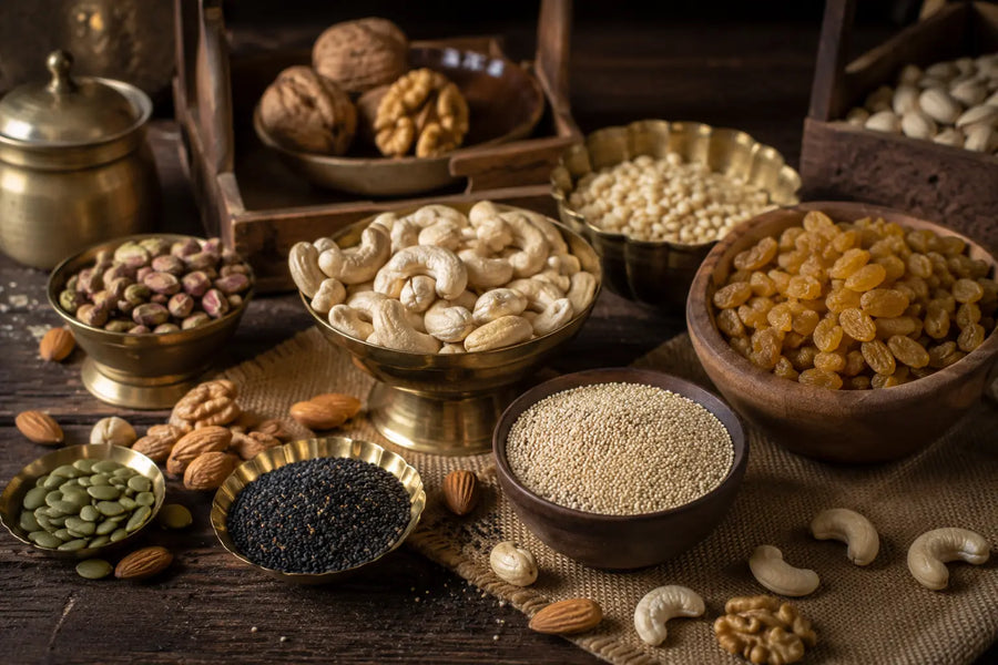 An assortment of whole nuts, seeds, and dried fruits arranged in brass and wooden bowls on a rustic dark wood table. Visible items include cashews, walnuts, almonds, pumpkin seeds, black seeds, sesame seeds, raisins, and pistachios.