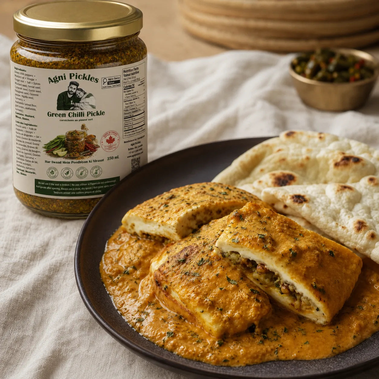 A jar of Agni Pickles Green Chilli Pickle is shown next to a plated dish of stuffed flatbreads in a rich orange-red sauce, set on a beige linen tablecloth.