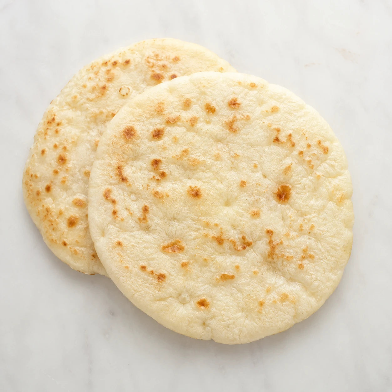 Two soft plain naan breads with golden blistered spots on a light neutral background.