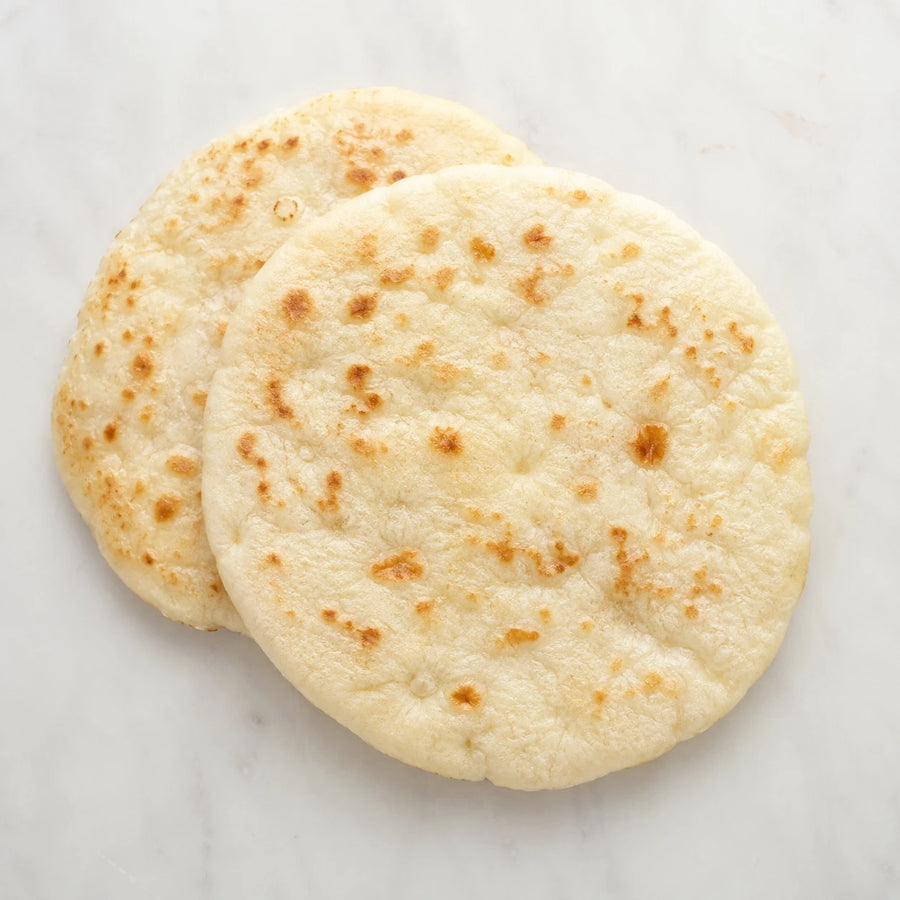 Two soft plain naan breads with golden blistered spots on a light neutral background.
