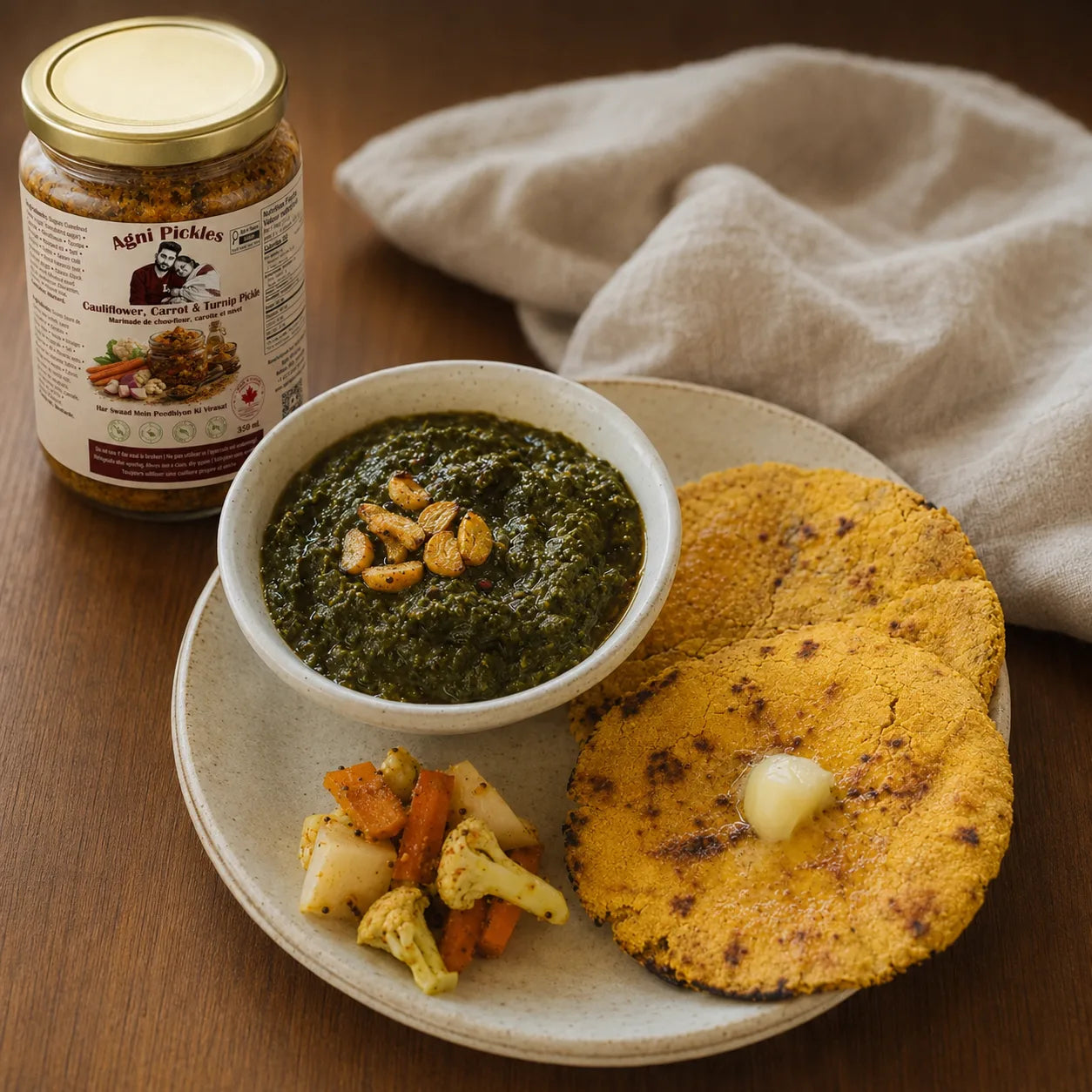 A jar of Mei Pickles Condiment and Spicy Sauce sits beside a plated meal on a wooden table. The plate holds a bowl of dark green chutney, two golden-brown fried flatbreads with butter, and small roasted vegetables, with a beige cloth in the background.