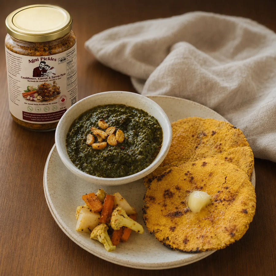 A jar of Mei Pickles Condiment and Spicy Sauce sits beside a plated meal on a wooden table. The plate holds a bowl of dark green chutney, two golden-brown fried flatbreads with butter, and small roasted vegetables, with a beige cloth in the background.