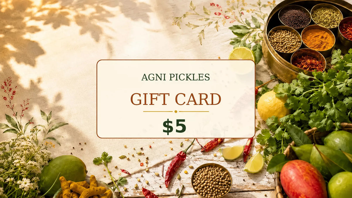 A 5 dollar Agni Pickles gift card centered on a rustic wooden table surrounded by fresh ingredients like limes, red chilies, coriander, ginger, mango, and a brass bowl of spices.
