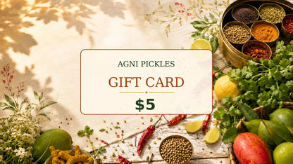A 5 dollar Agni Pickles gift card centered on a rustic wooden table surrounded by fresh ingredients like limes, red chilies, coriander, ginger, mango, and a brass bowl of spices.