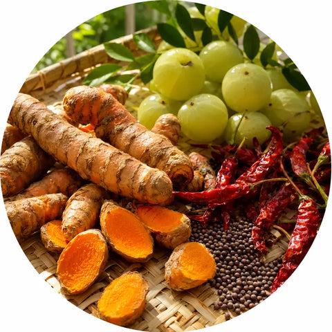 A circular close-up of fresh turmeric roots, sliced orange turmeric pieces, green grapes, dried red chilies, and dark seeds arranged on a woven tray.