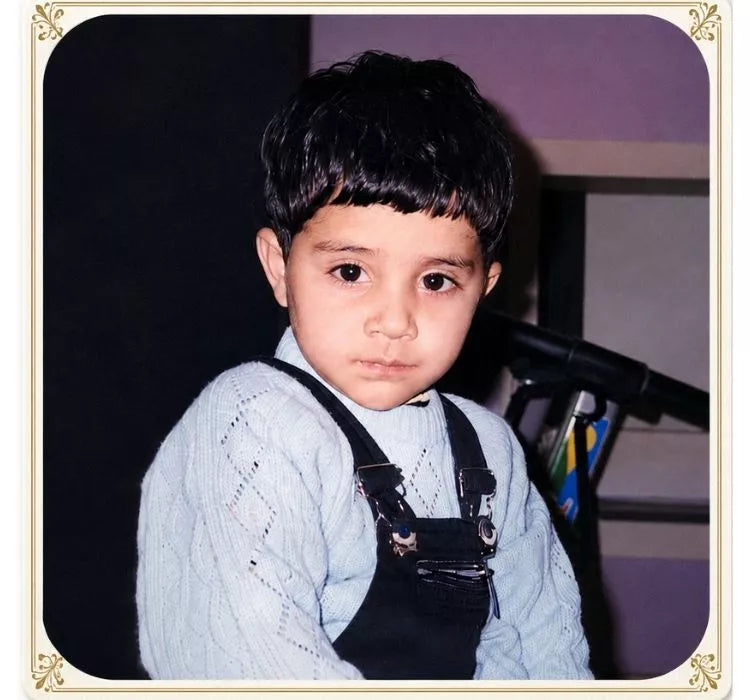 Young child wearing a light blue sweater and black overalls sitting in front of a dark background.