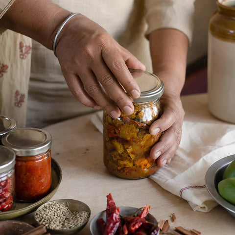 A person's hands are shown sealing a glass jar filled with pickled or fermented vegetables on a rustic wooden table surrounded by other jars of spices and ingredients.