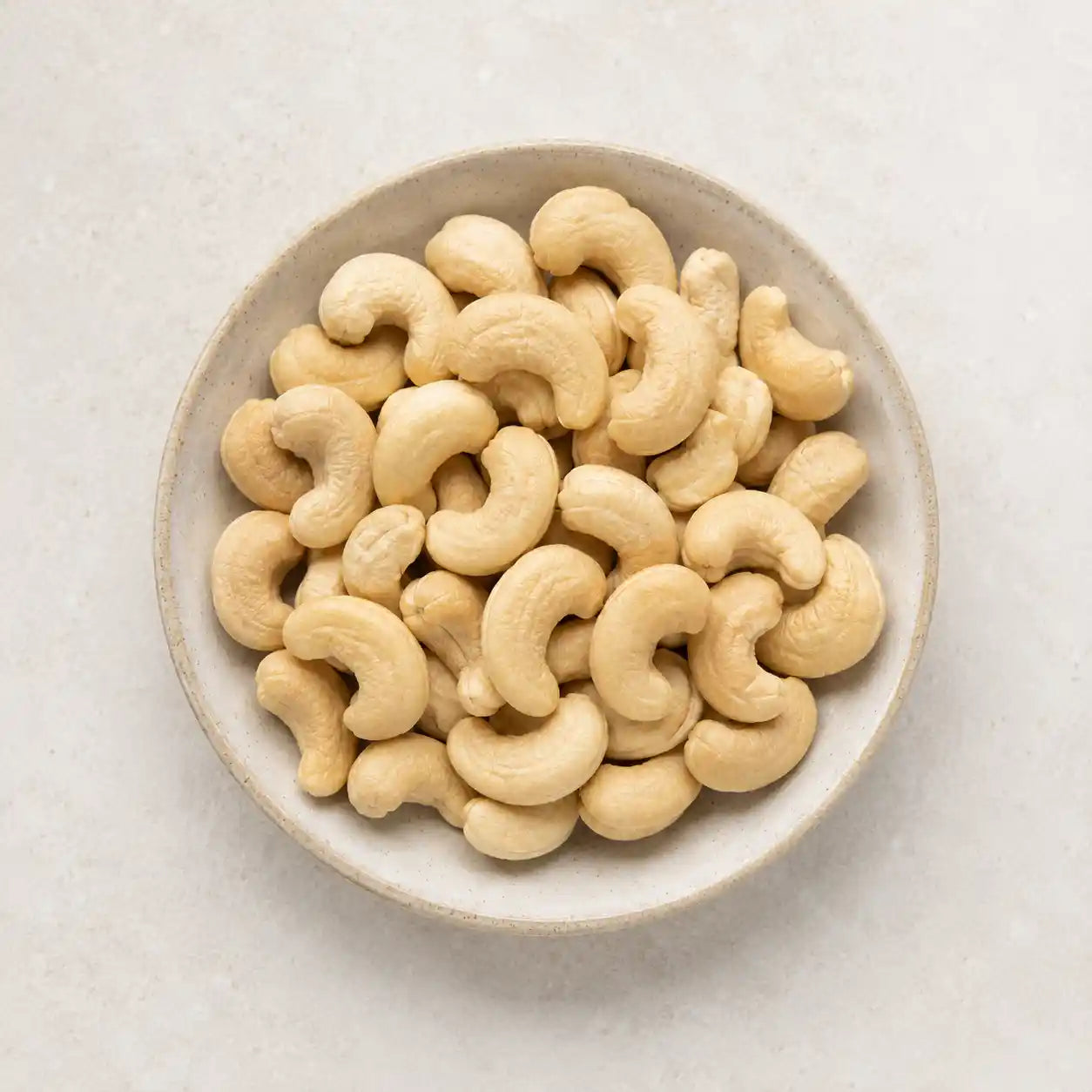 Raw cashews piled in a round off-white ceramic bowl on a light beige textured surface.