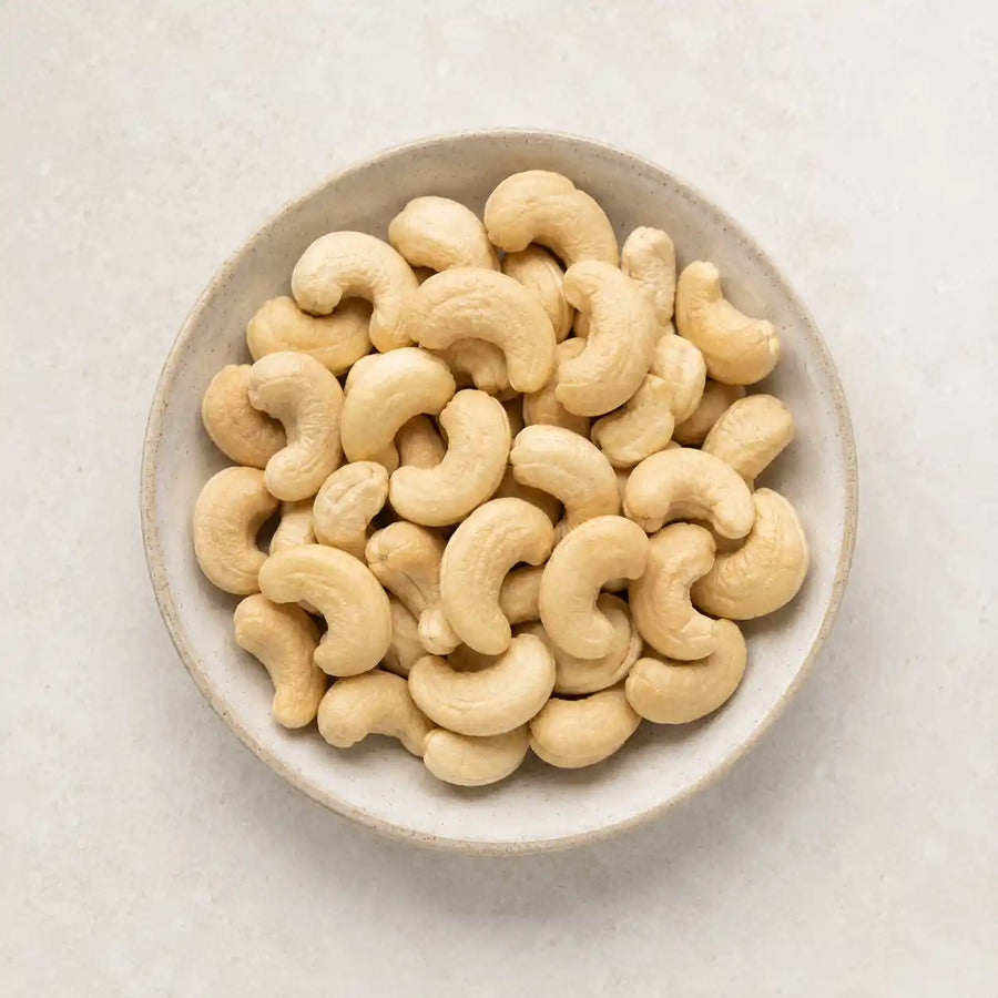 Raw cashews piled in a round off-white ceramic bowl on a light beige textured surface.