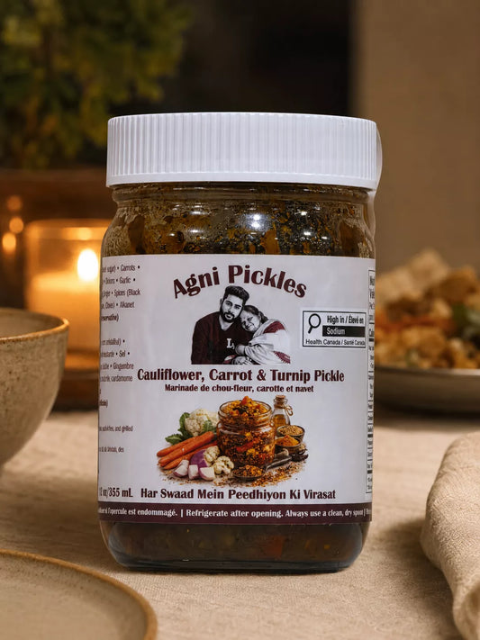 A jar of Agni Pickles Cauliflower, Carrot & Turnip Pickle, set on a beige tablecloth with warm, softly blurred background elements including a lit candle and food, indicating a lifestyle or styled environment.