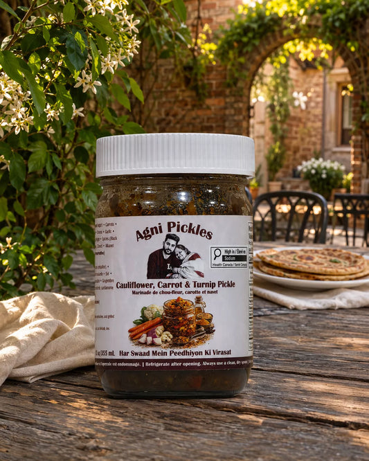 A jar of Agni Pickles Cauliflower, Carrot & Turnip Pickle sits on a rustic wooden table outdoors. The background features a warm, sunlit patio setting with a stone archway, green ivy, and a stack of flatbreads on a tablecloth.