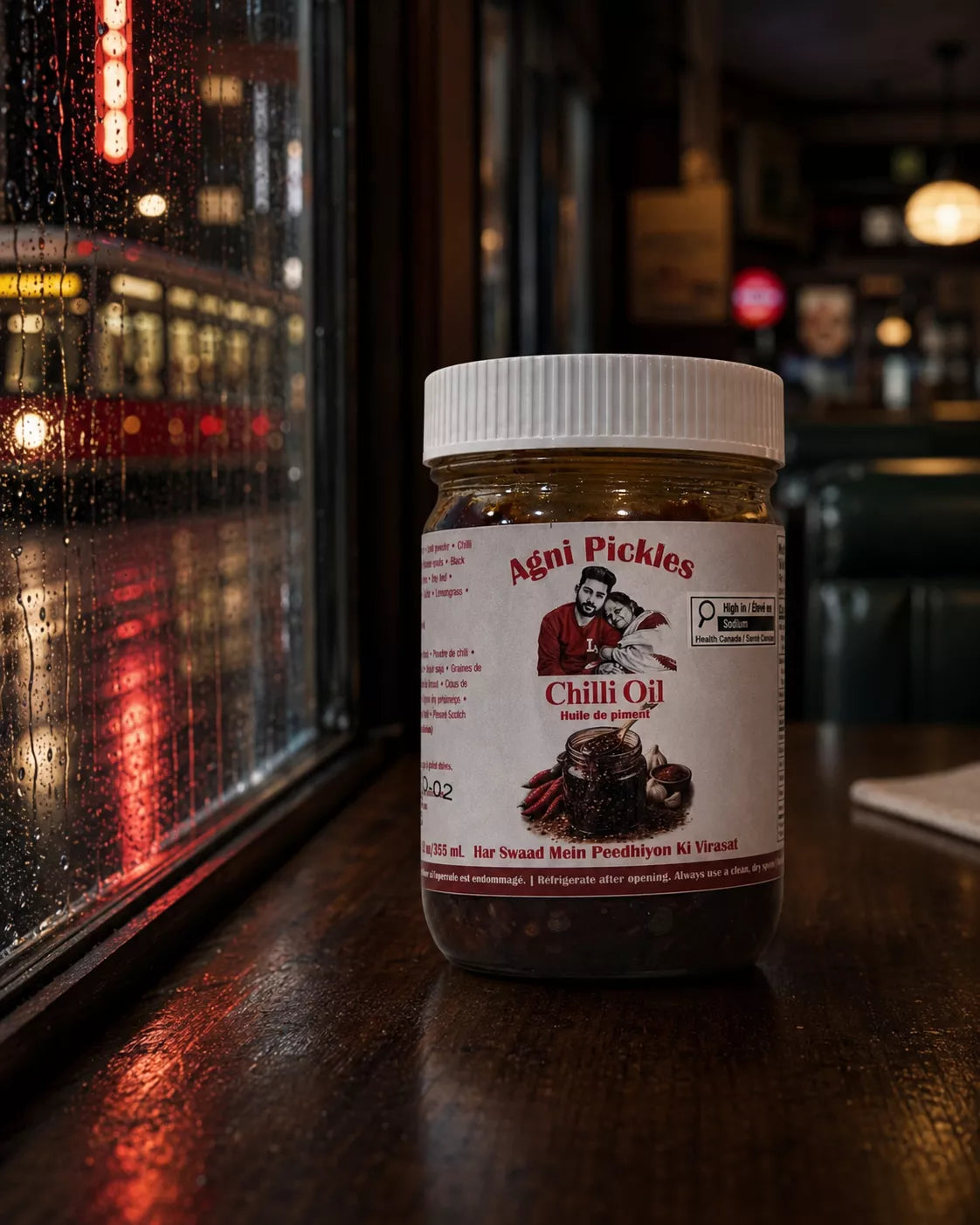 A jar of Aéni Pickles Chili Oil sits on a dark wooden table in a dimly lit restaurant or bar setting, with a rainy window and blurred lights in the background.