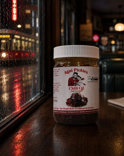 A jar of Aéni Pickles Chili Oil sits on a dark wooden table in a dimly lit restaurant or bar setting, with a rainy window and blurred lights in the background.