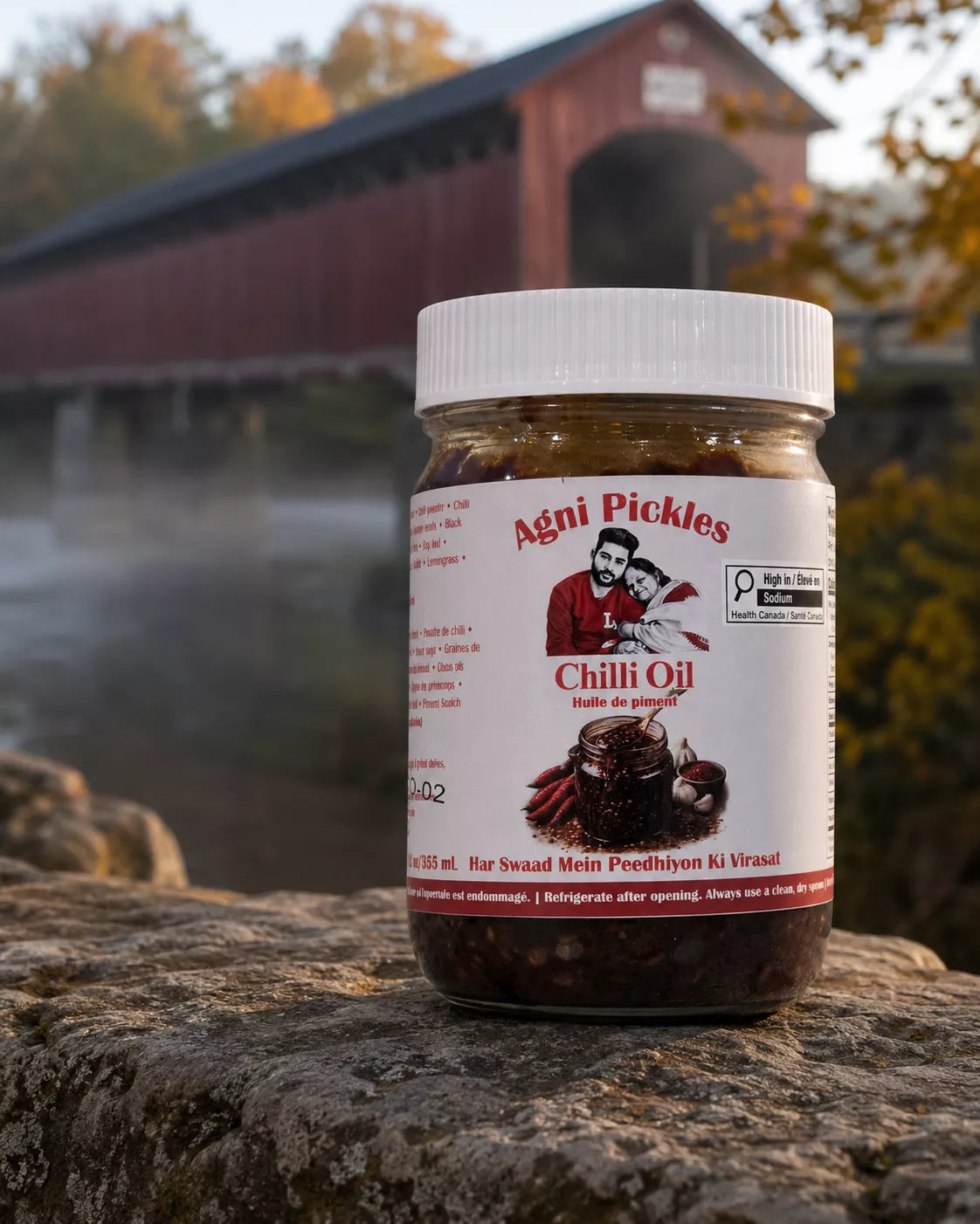 A jar of Agni Pickles Chilli Oil placed on a stone surface with a blurred red covered bridge and autumn trees in the background.