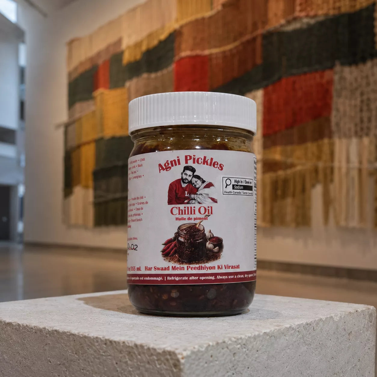 A jar of Agni Pickles Chili Oil placed on a light stone pedestal, with a blurred multicolored textile background.
