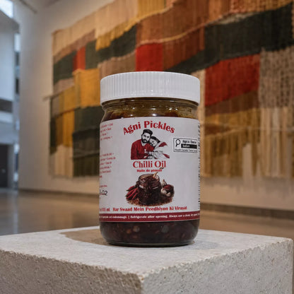 A jar of Agni Pickles Chili Oil placed on a light stone pedestal, with a blurred multicolored textile background.