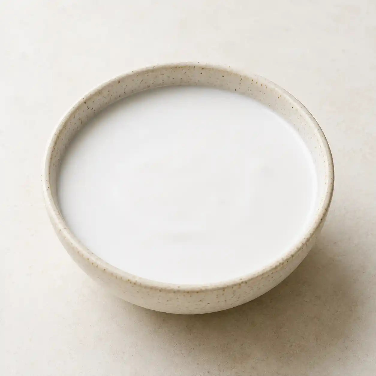 A small speckled ceramic bowl filled with white yogurt, photographed from a top-down angle on a light beige textured surface.