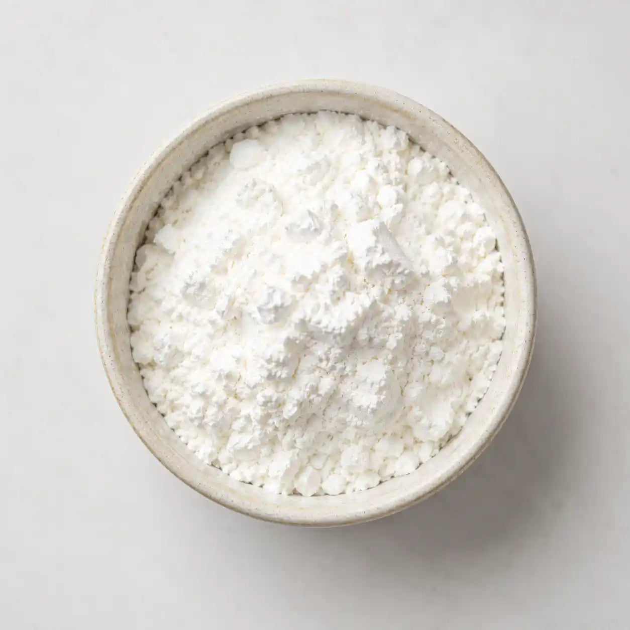 A top-down view of a white powder, likely flour or starch, piled in a small beige ceramic bowl against a light gray background.