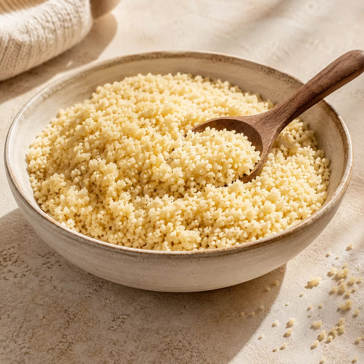 A close-up of cooked couscous in a beige ceramic bowl with a wooden spoon, set on a textured beige surface with a soft cloth in the background.