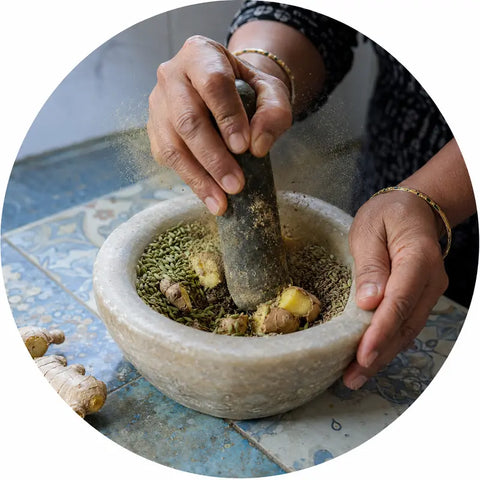 A close-up circular photo of a person's hands using a stone pestle to grind herbs and ginger root inside a large stone mortar on a patterned blue tile surface.