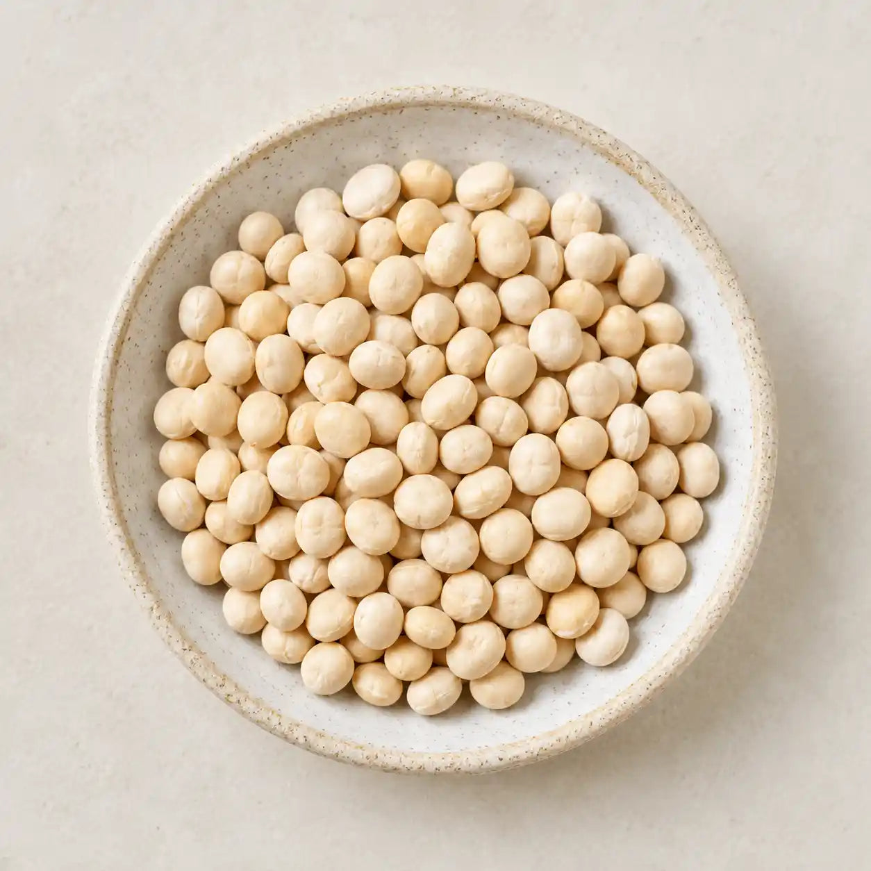 A top-down view of light beige soybeans piled in a round speckled ceramic bowl on a light beige surface.