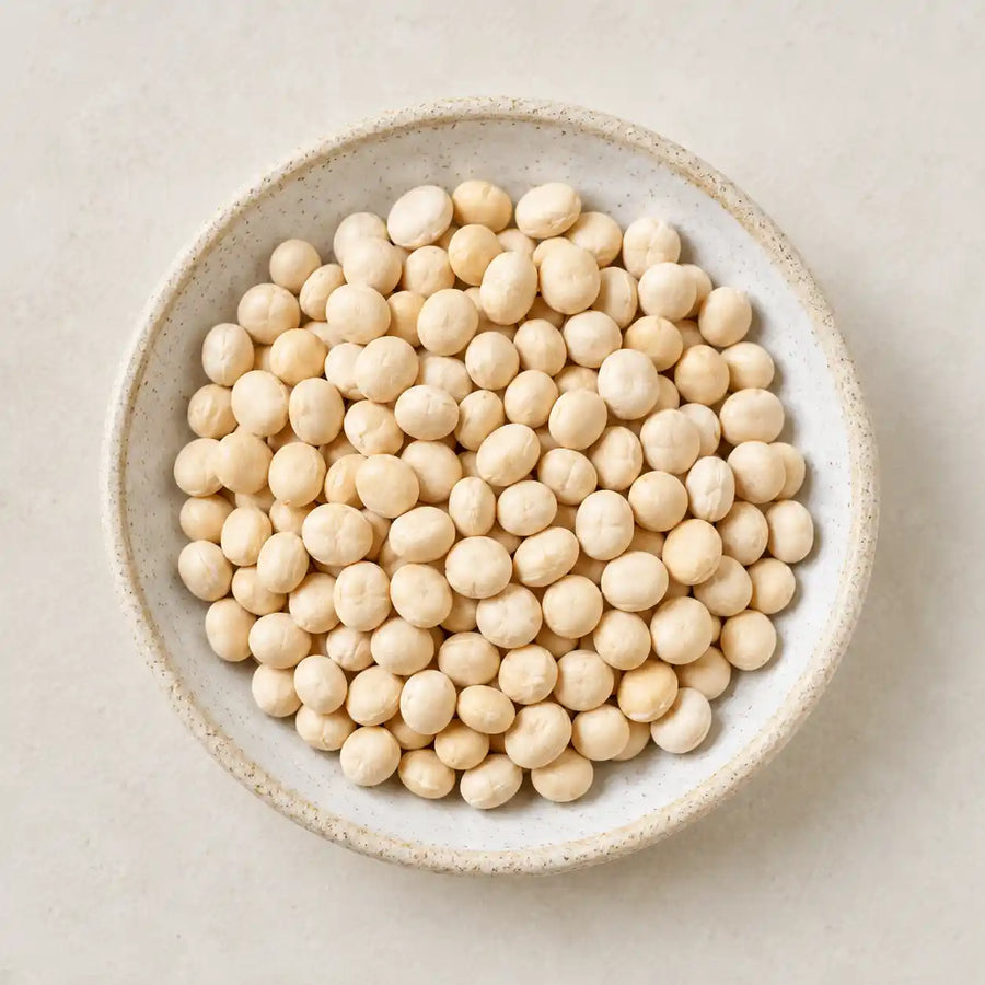 A top-down view of light beige soybeans piled in a round speckled ceramic bowl on a light beige surface.