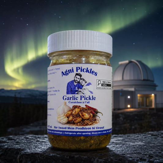 A jar of Agni Pickles Garlic Pickle stands on a dark stone surface with a blurred night sky background featuring green aurora borealis and an observatory dome. The product jar has a white label and white cap, showing yellowish-green pickled contents.