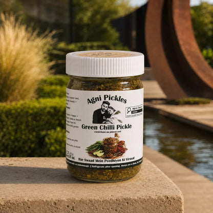 A jar of Agni Pickles Green Chilli Pickle sits on a stone ledge outdoors, with a garden, water feature, and rusted metal sculpture in the background. The product is a clear glass jar with a white ribbed screw cap, showing the green pickled contents, and a white label with the Agni Pickles brand name and product details.