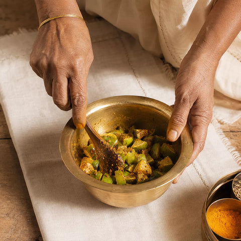 A close-up of a person's hands mixing a brass bowl of cooked green vegetables and spices on a cream-colored textile.