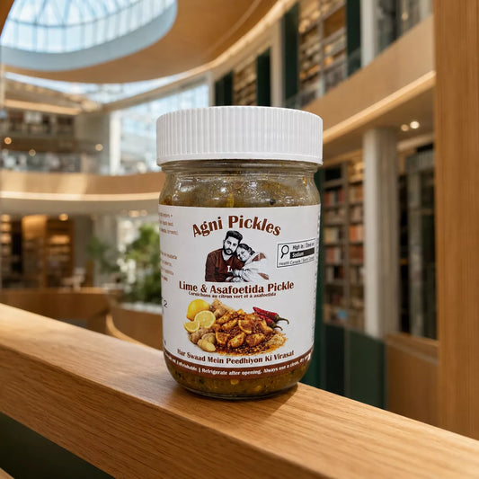 A jar of Agni Pickles Lime & Asafoetida Pickle sits on a wooden railing in a modern, sunlit library or bookstore interior.