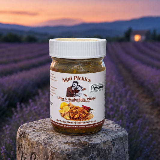 A jar of Agni Pickles Lime & Asafoetida Pickle sits on a stone pillar in a lavender field at sunset. The jar has a white lid and a white label with red and black text, showing a picture of a man and a piece of chicken.