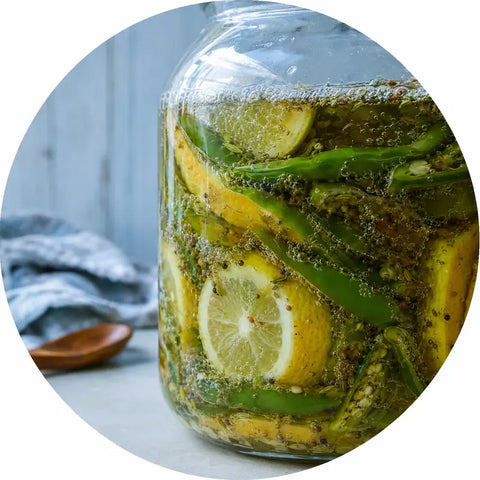A large glass jar filled with lemon slices, green beans, and peppercorns submerged in a brine, shown in a circular frame with a rustic kitchen background.