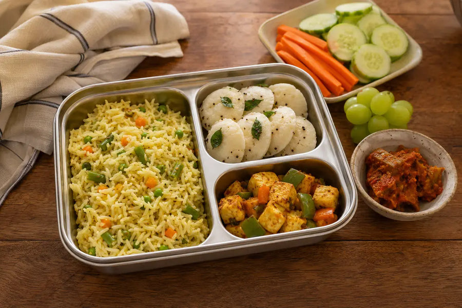 Vegetarian Indian lunchbox with vegetable pulao, mini idlis, paneer bites, fresh vegetables, grapes and pickle.