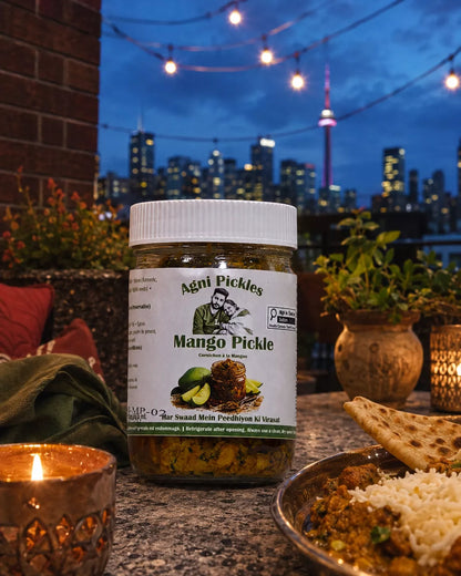 A jar of Agni Pickles Mango Pickle is displayed on a stone table in a restaurant setting with a city skyline at dusk in the background. A lit candle, a plate of food, and potted plants are visible around the jar.