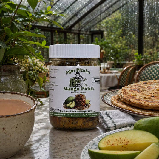 A jar of Agni Pickles Mango Pickle sits on a table in a greenhouse or garden setting, surrounded by fresh mango slices, a cup of tea, and flatbread on a plate.