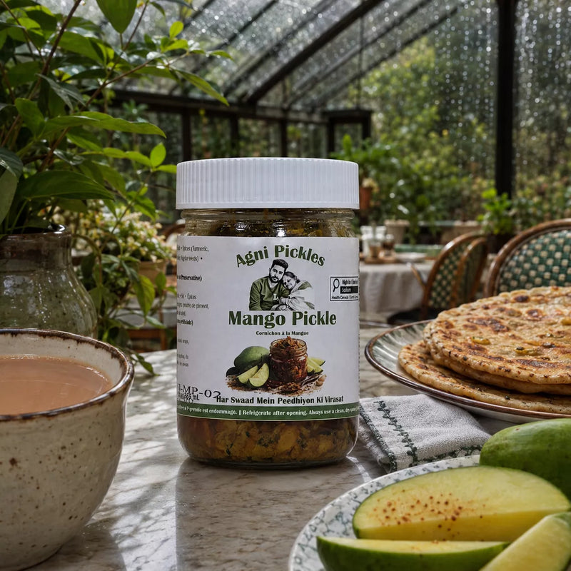 A jar of Agni Pickles Mango Pickle sits on a table in a greenhouse or garden setting, surrounded by fresh mango slices, a cup of tea, and flatbread on a plate.