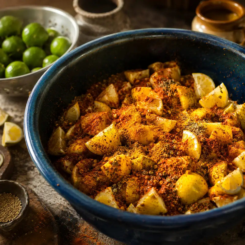 Cut lemons layered with turmeric, chili powder, salt, and whole spices in a blue mixing bowl.